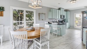 A modern kitchen with white cabinetry, a central island, and a dining area with white chairs. - Wonderful Ocean Views - Sleeps 12 -3 Mins to Beach (Oak Island)