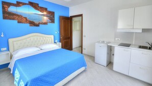 Une chambre à coucher avec un lit, un mur bleu, un tableau, une porte et un coin cuisine avec des placards et un évier.