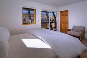 A bedroom with a bed, a chair, and two windows with views of a landscape.