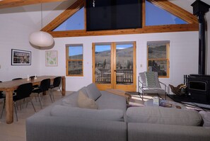 Una sala de estar acogedora con techo alto, un comedor con mesa y sillas de madera, y ventanas amplias con vistas a un paisaje montañoso.