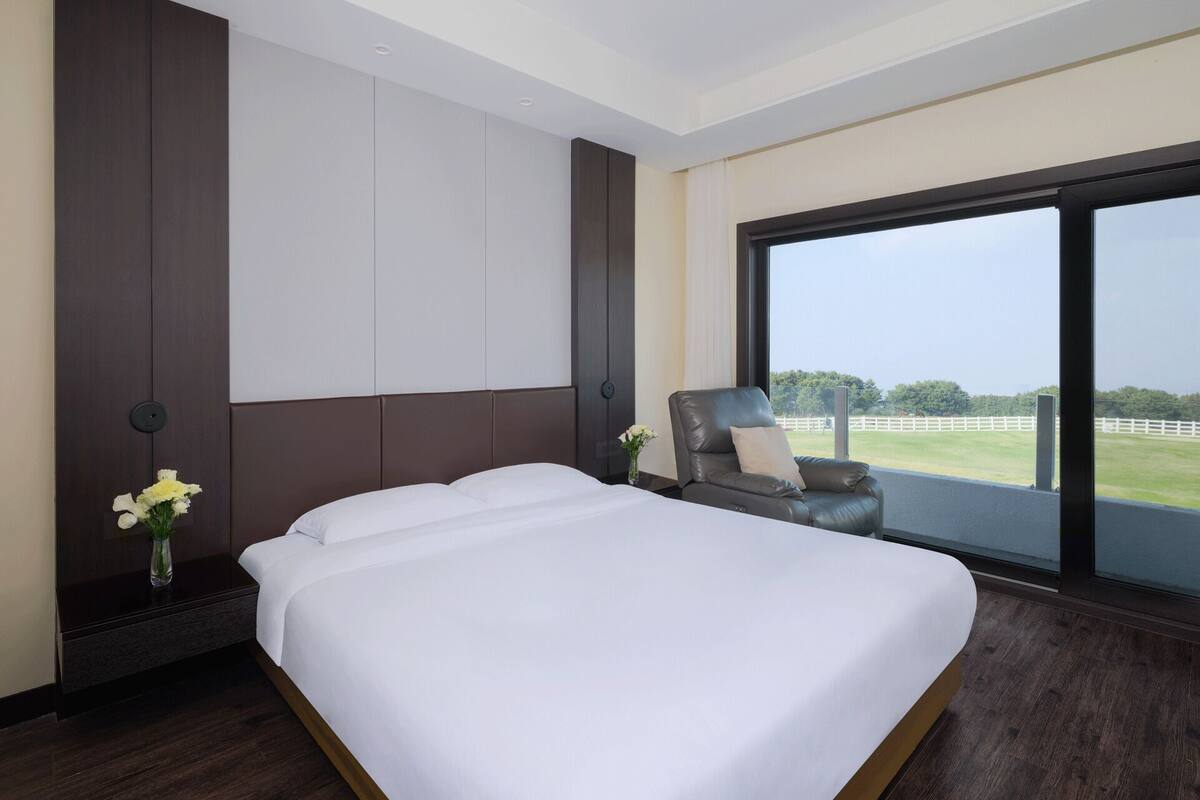 A modern hotel room with a large bed, a seating area with a chair, a nightstand, and a large window offering a view of the outdoors.