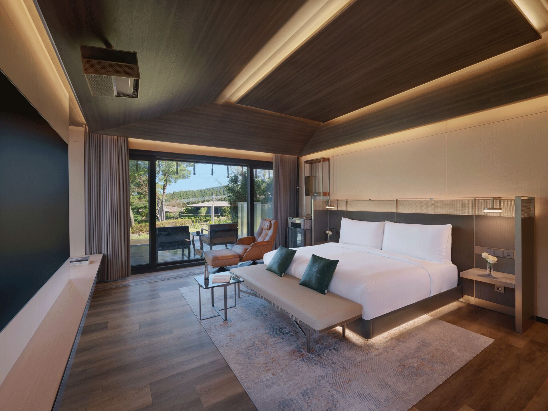 A modern bedroom with a large bed, a bench, a desk, and a view of the outdoors.