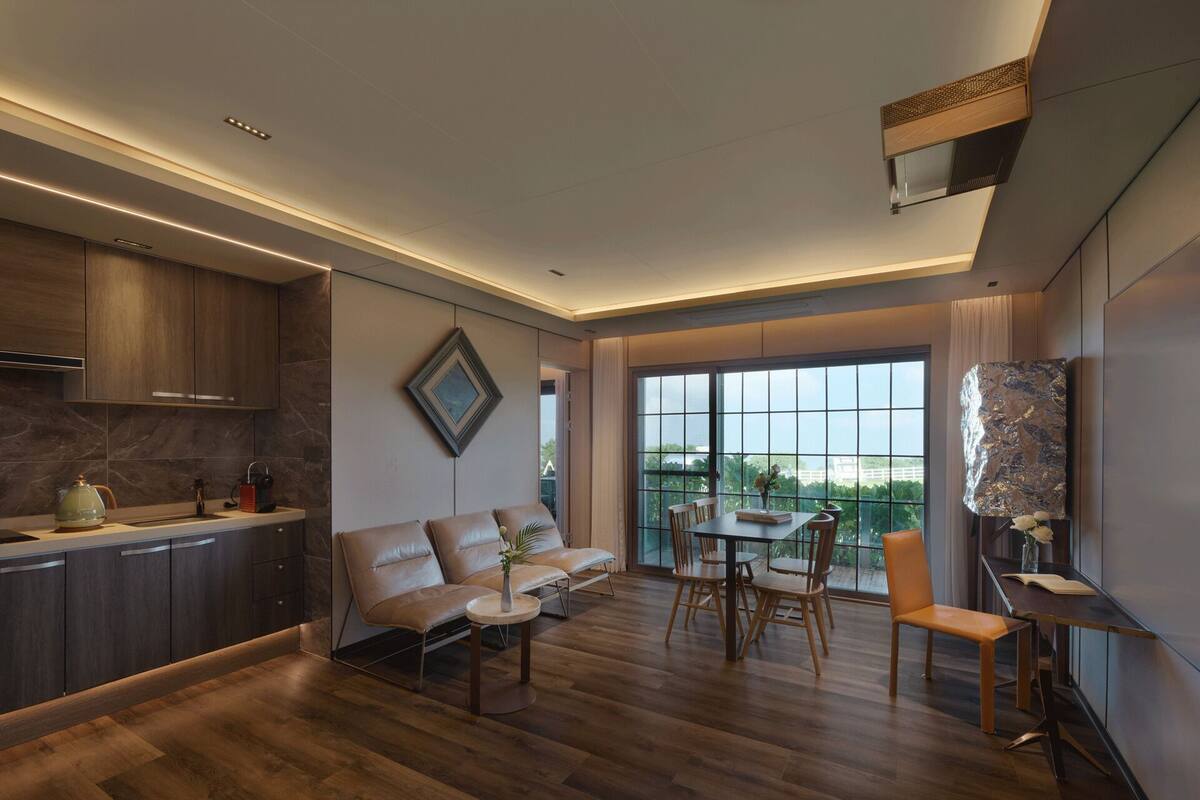 A modern kitchen with dark wood cabinets, a dining area with a wooden table and chairs, and a large window offering a view of greenery.