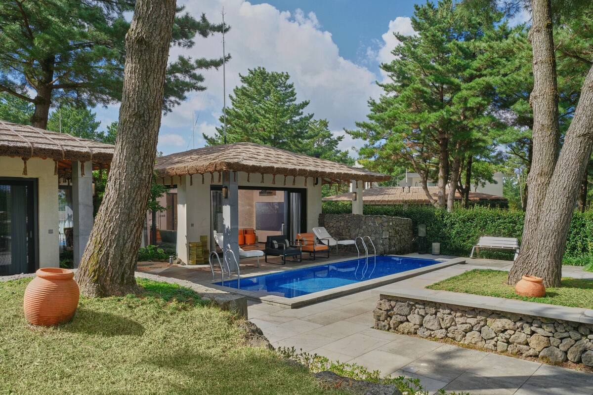 A pool area with a stone wall and lounge chairs, surrounded by trees and a building with a tiled roof.