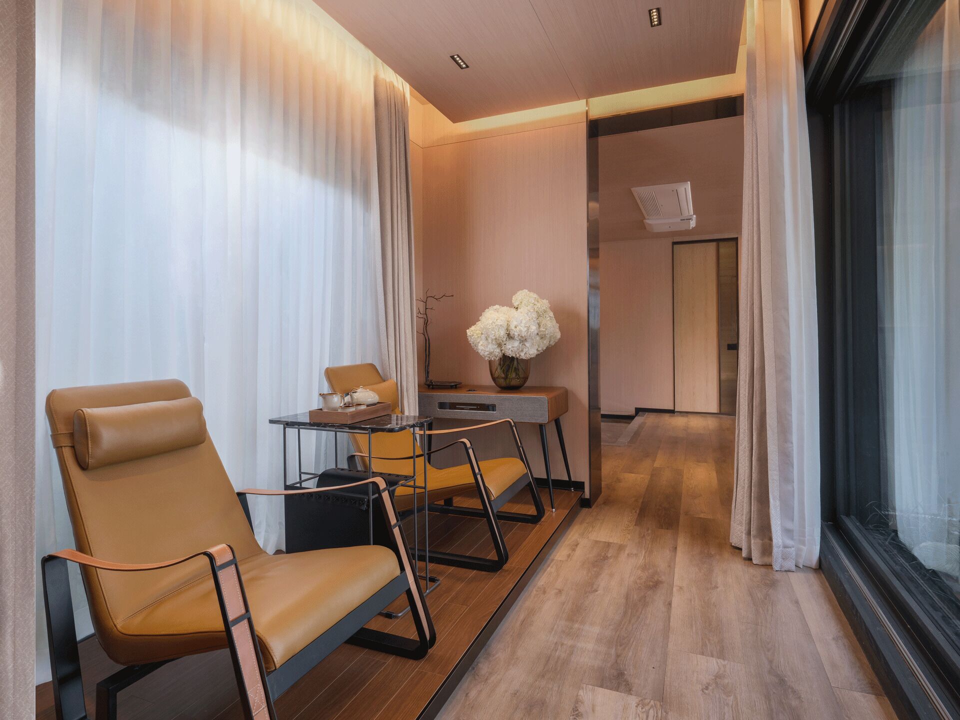 A modern hotel room with a seating area featuring two leather chairs, a small table with a vase of flowers, and a view through a large window.