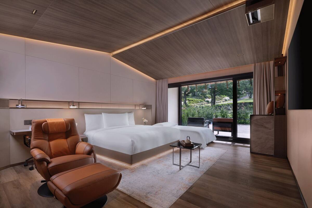 A modern hotel room with a large bed, a leather chair, a small table, and a view of the outdoors.