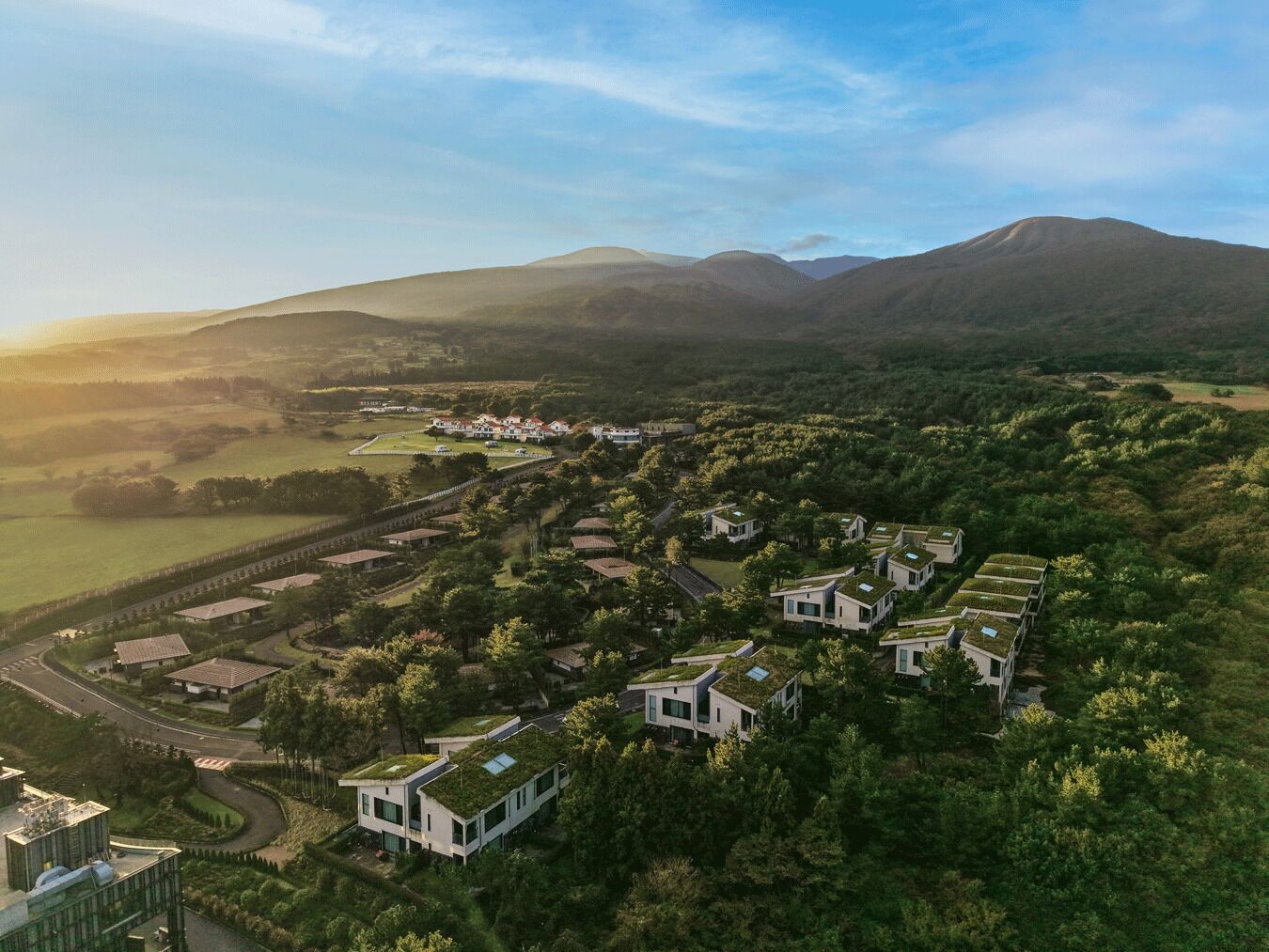 A residential area with modern houses featuring green roofs, surrounded by lush greenery and hills in the background.