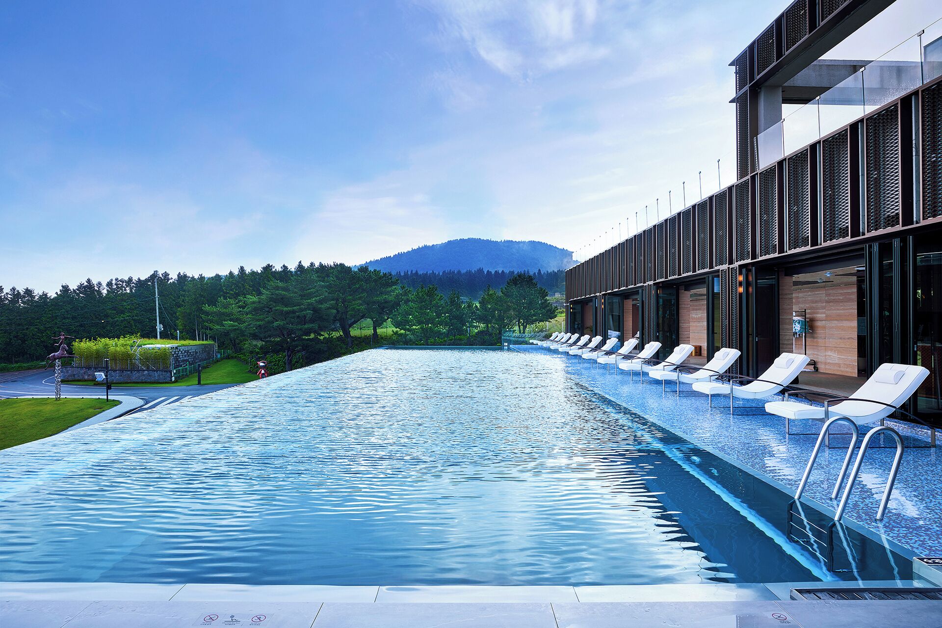 A modern hotel with a long swimming pool, lounge chairs, and a view of a forested mountain range.