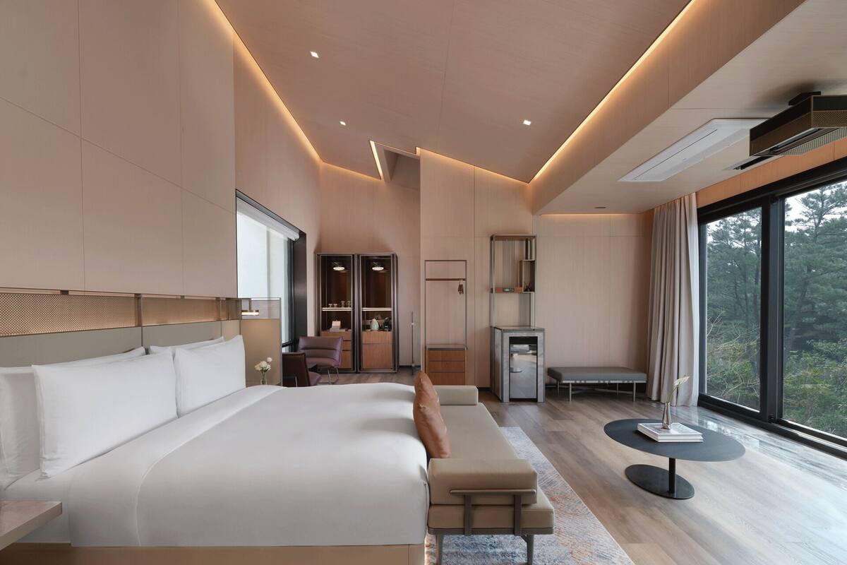 A modern hotel room with a large bed, a seating area, a small table, and a view of the outdoors.