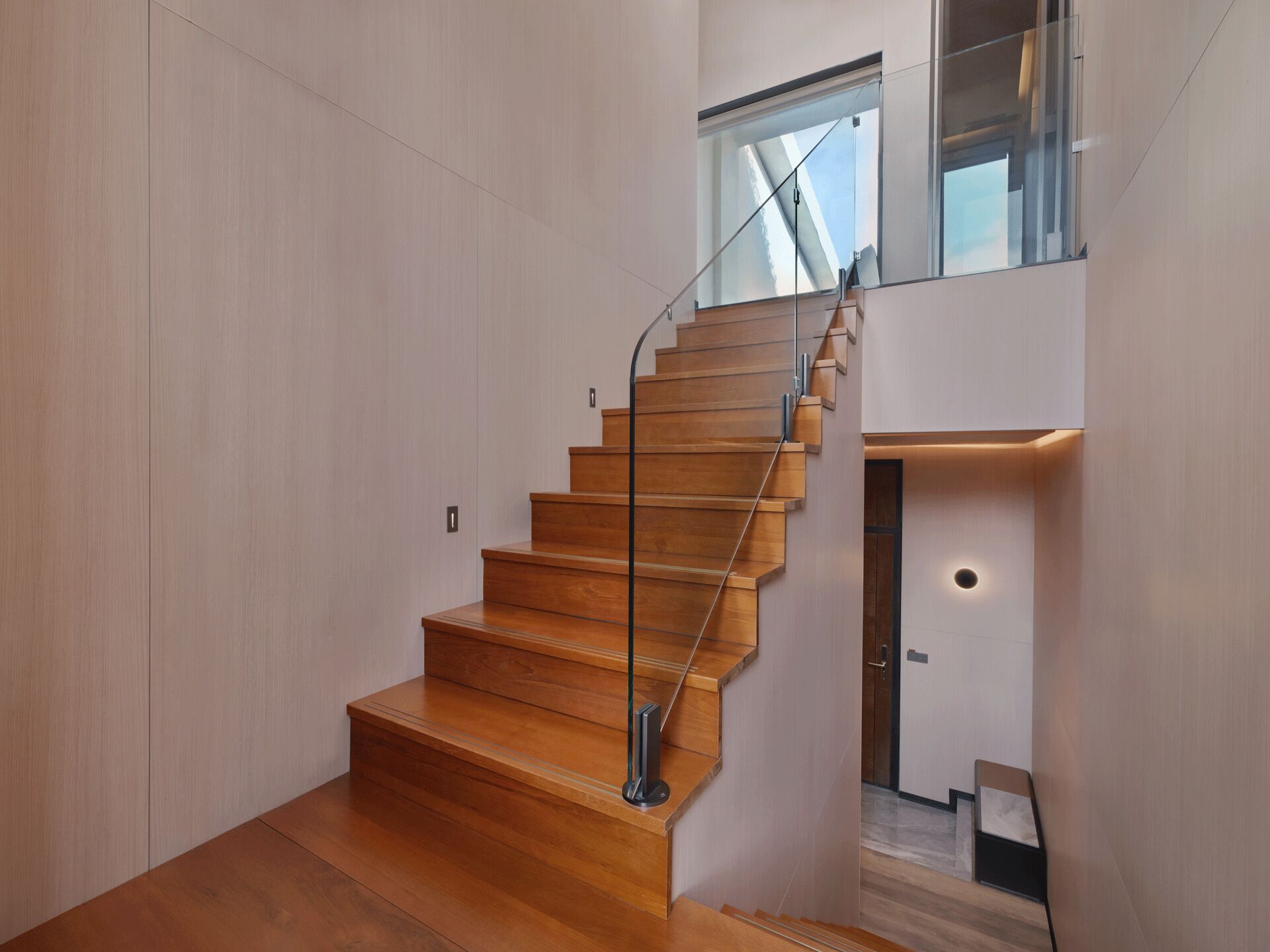 A modern staircase with wooden steps and glass railings, leading to a second-floor landing with a door and a small table.
