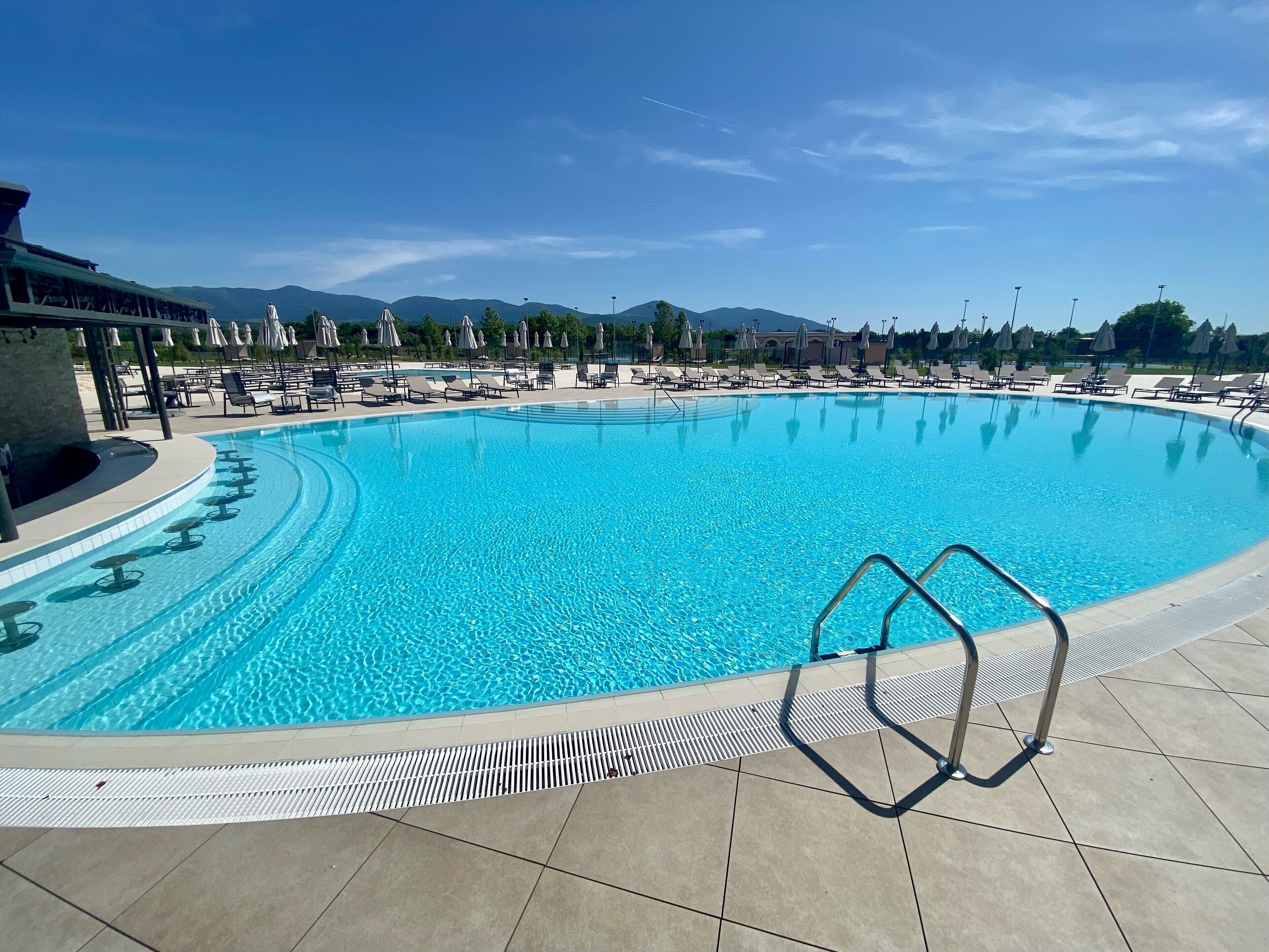 Una piscina amplia y cristalina con sillas de descanso y sombrillas, rodeada de un solárium con baldosas y montañas al fondo.