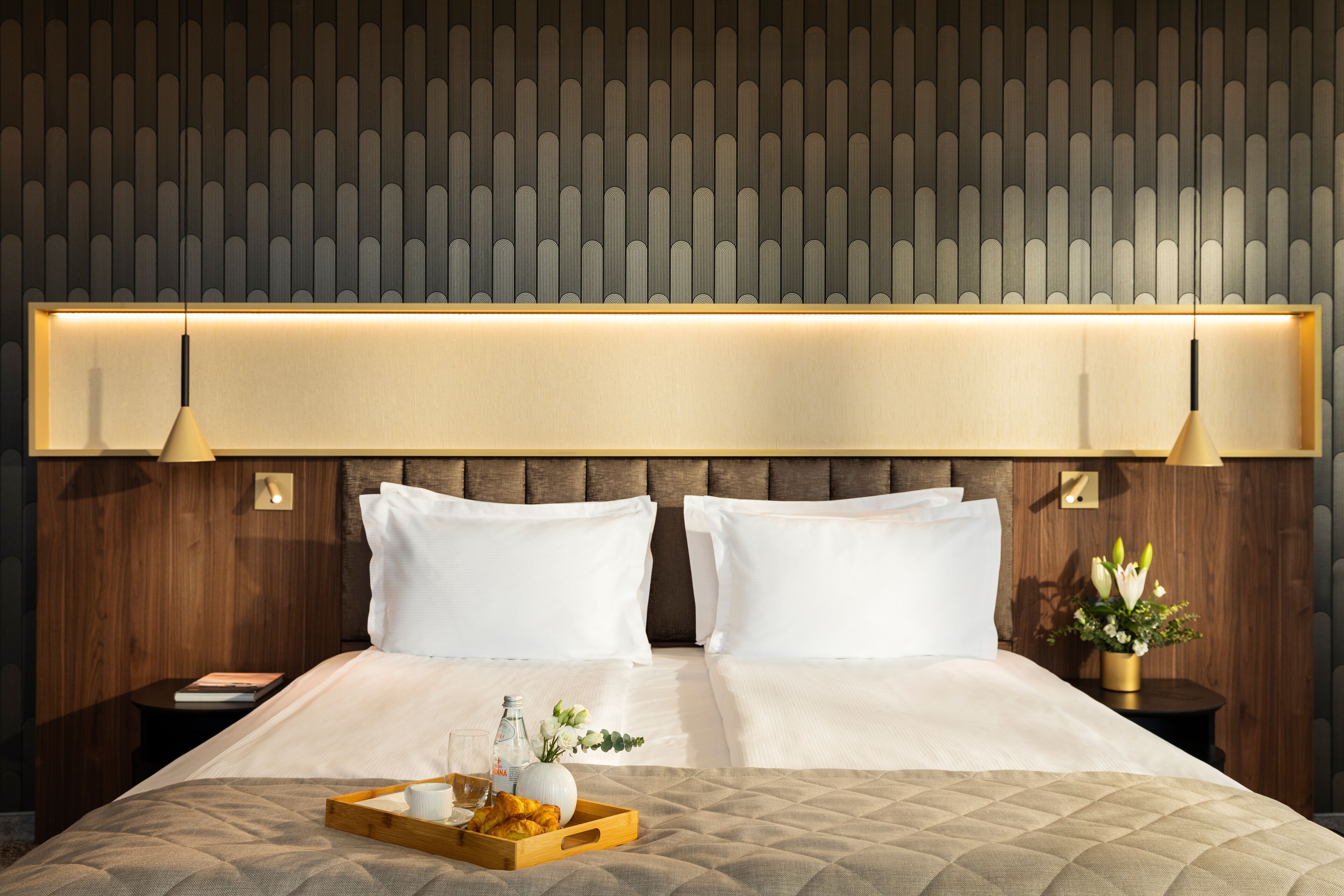 A hotel room with a large bed, two white pillows, a wooden headboard, a bedside table with a lamp, and a tray with a cup and flowers.