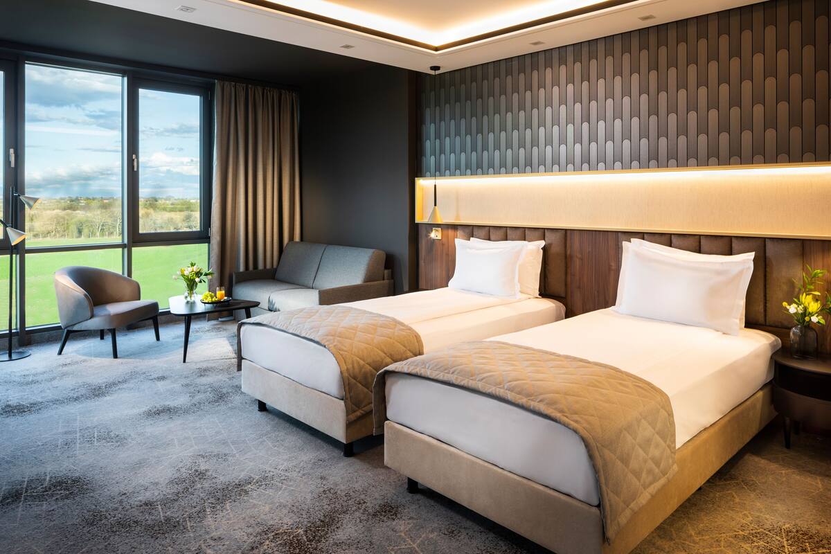 A hotel room with two beds, a sofa, a small table with a vase of flowers, and a view of the outdoors.