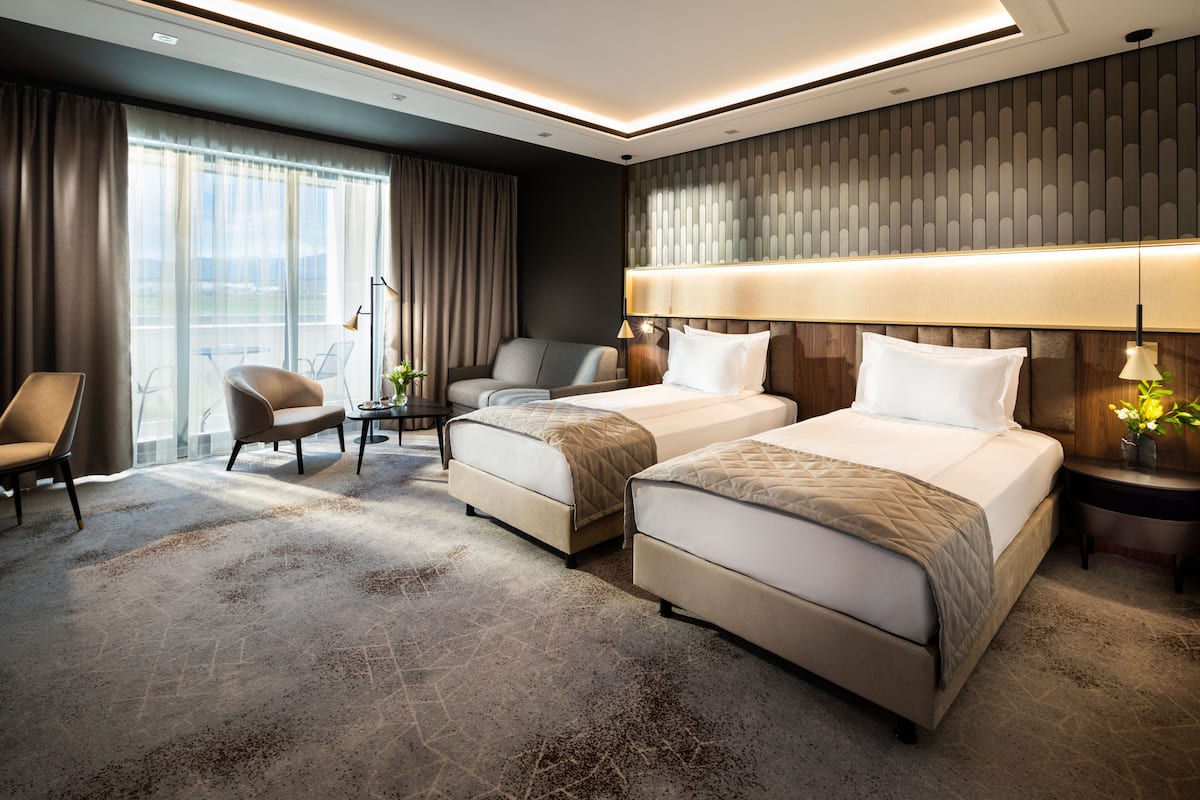 A hotel room with two beds, a desk, a chair, and a large window with curtains.