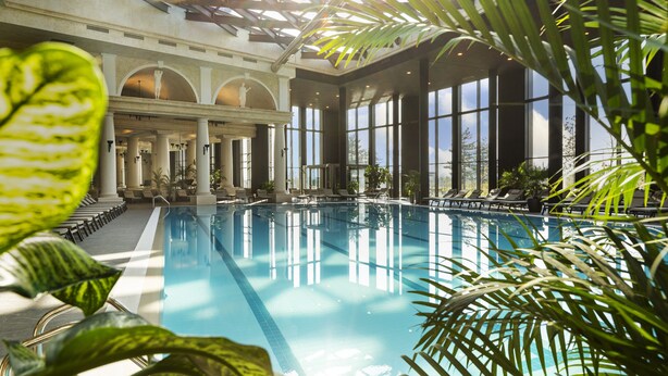 An indoor swimming pool with clear water, surrounded by large windows and columns, and lush green plants. - Kings Valley Medical & Spa Hotel (Kazanlak)