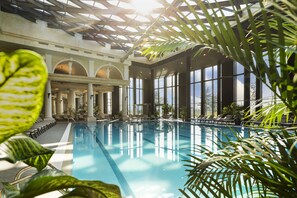 An indoor swimming pool with clear water, surrounded by large windows and columns, and lush green plants. - Kings Valley Medical & Spa Hotel (Kazanlak)