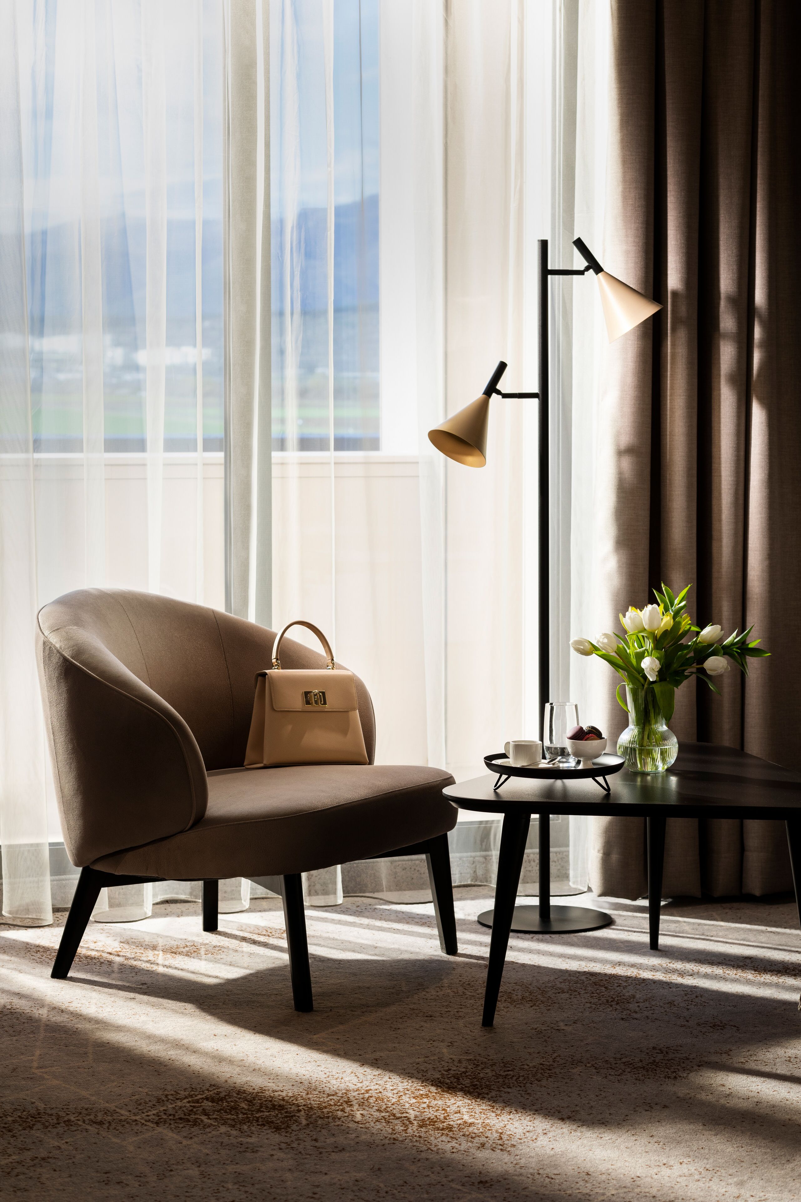 Una habitación moderna con un sillón de cuero, una lámpara de pie, una mesita con un jarrón de flores y vistas al exterior a través de cortinas traslúcidas.