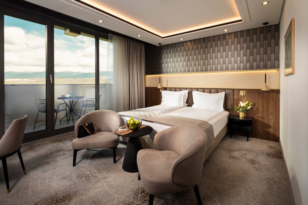 A modern hotel room with a large bed, two armchairs, a small table with a bowl of fruit, and a balcony with a table and chairs.