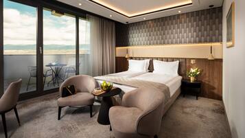 Quarto de hotel moderno com uma cama grande, duas poltronas, uma mesinha de centro com uma cesta de frutas e uma varanda com mesa e cadeiras.