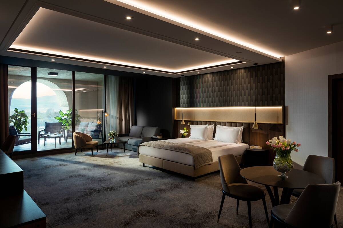 A modern hotel room with a large bed, a sitting area with a sofa and chairs, a dining table, and a view through large windows.
