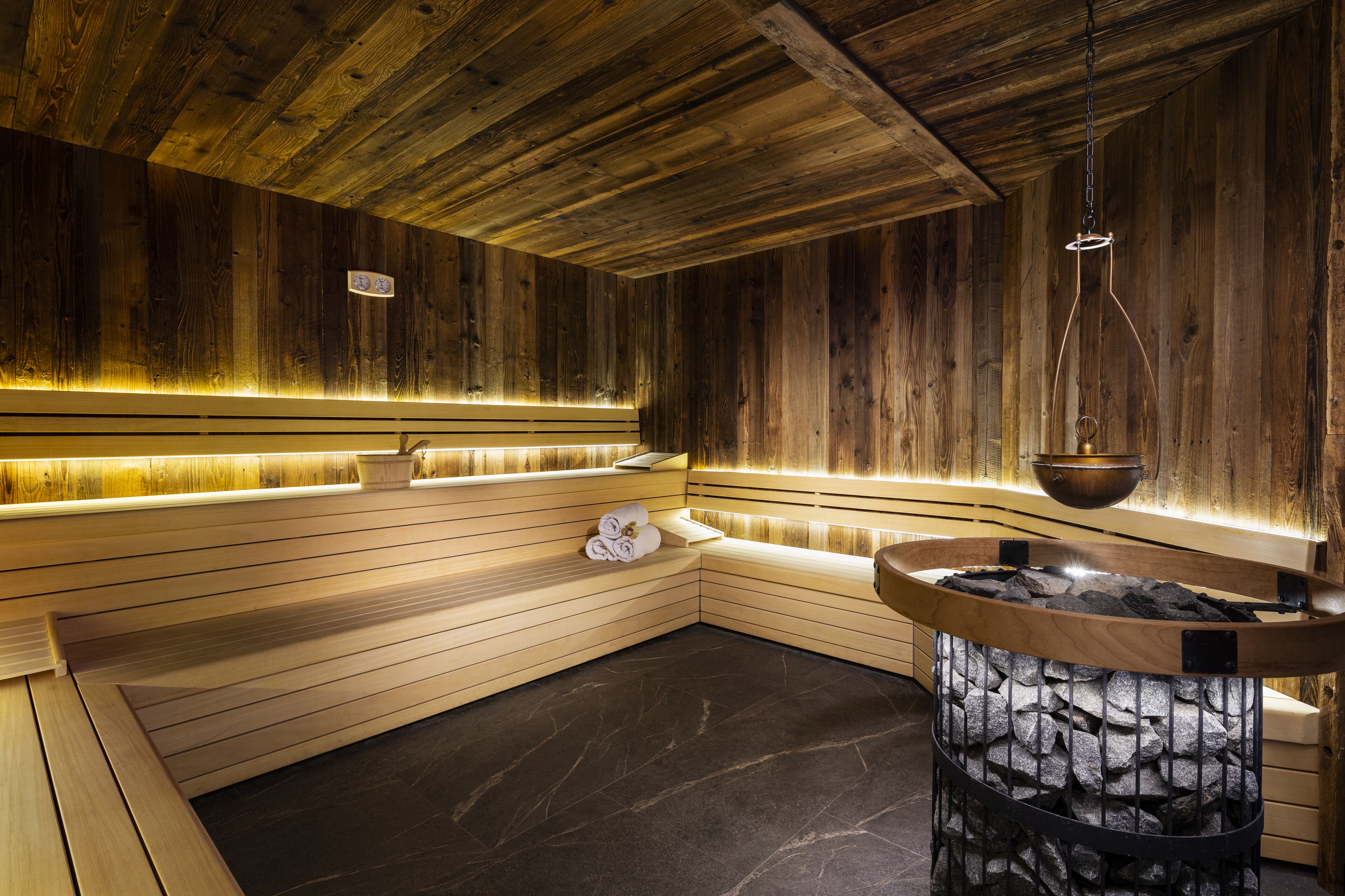 Un sauna aux murs et aux bancs en bois, doté d’un sol en pierre et d’un luminaire suspendu.