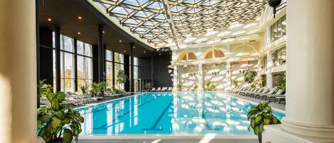 An indoor swimming pool with lounge areas, large windows, and a high ceiling with a geometric glass design.