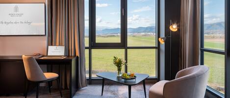 Een moderne hotelkamer met een bureau, twee stoelen, een kleine tafel met een bloevas en een schaal fruit, en een groot raam met uitzicht op een groen veld en bergen.