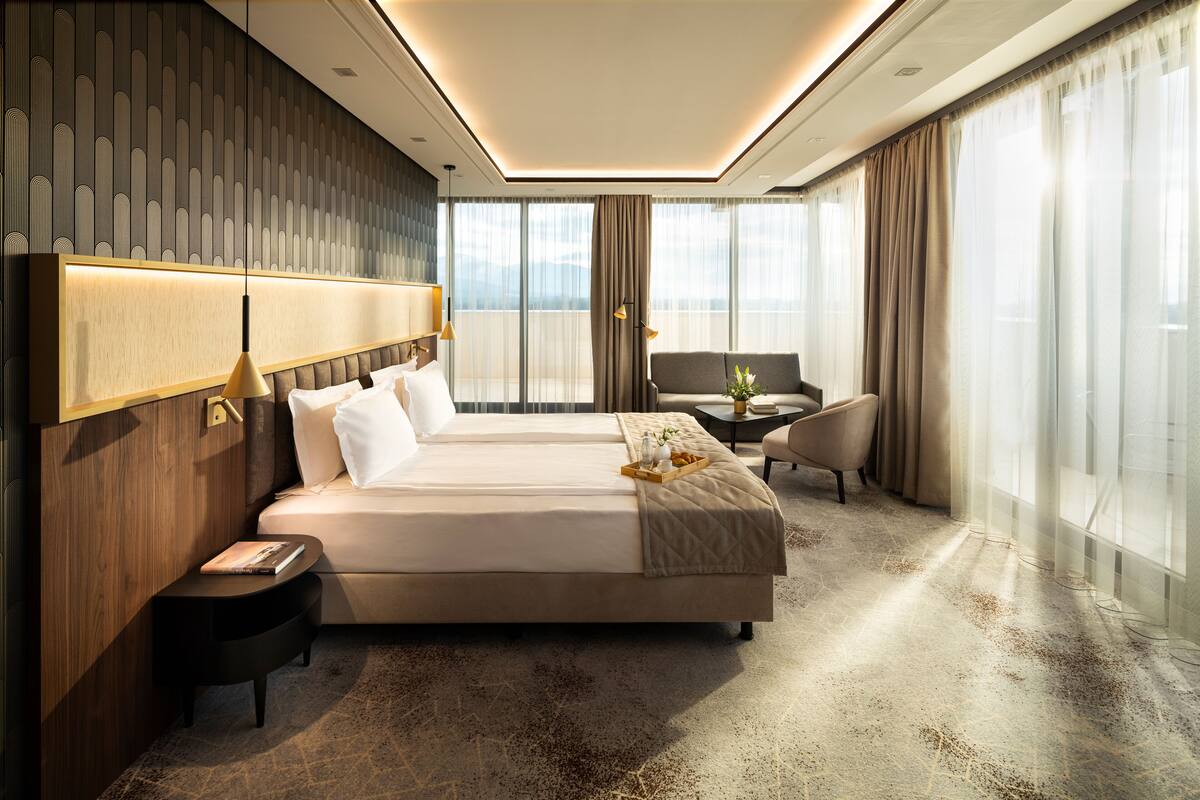A modern hotel room with a large bed, a small sofa, a side table, and a view of the city through large windows.