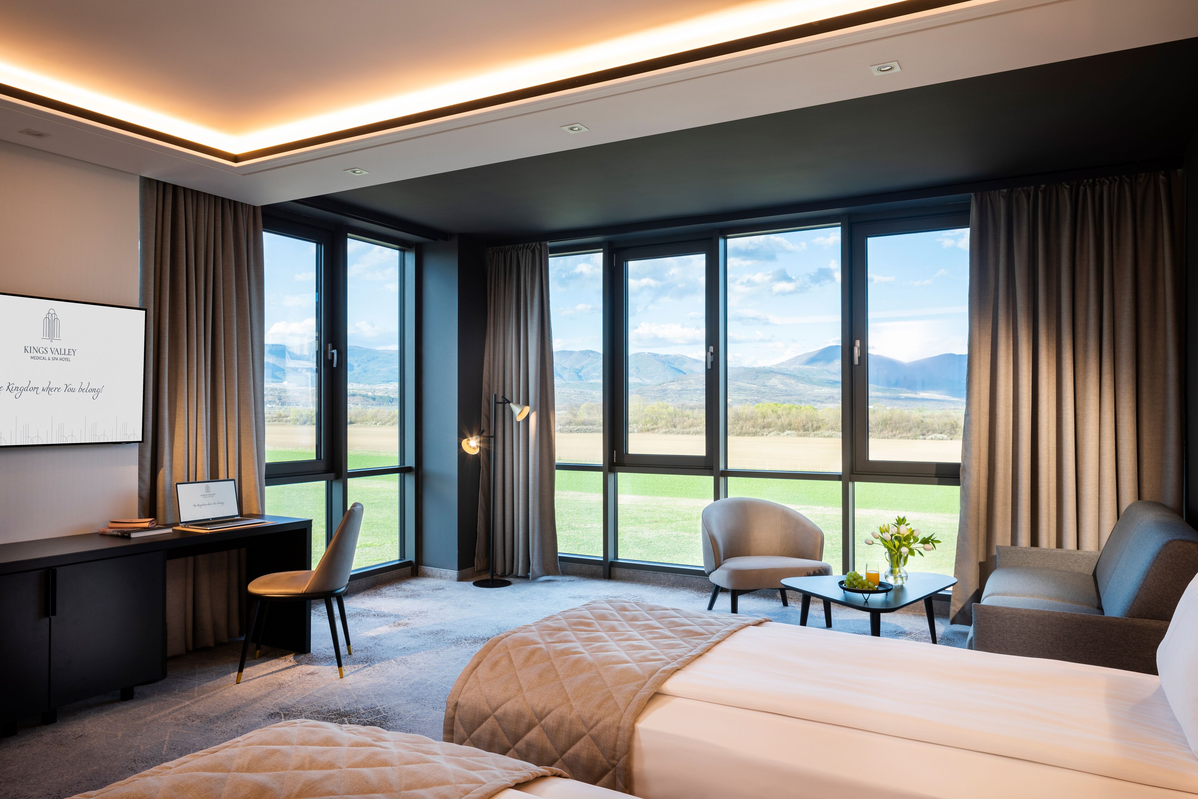 A hotel room with a large bed, a desk with a laptop, two chairs, a small table with a vase, and large windows offering a mountain view.