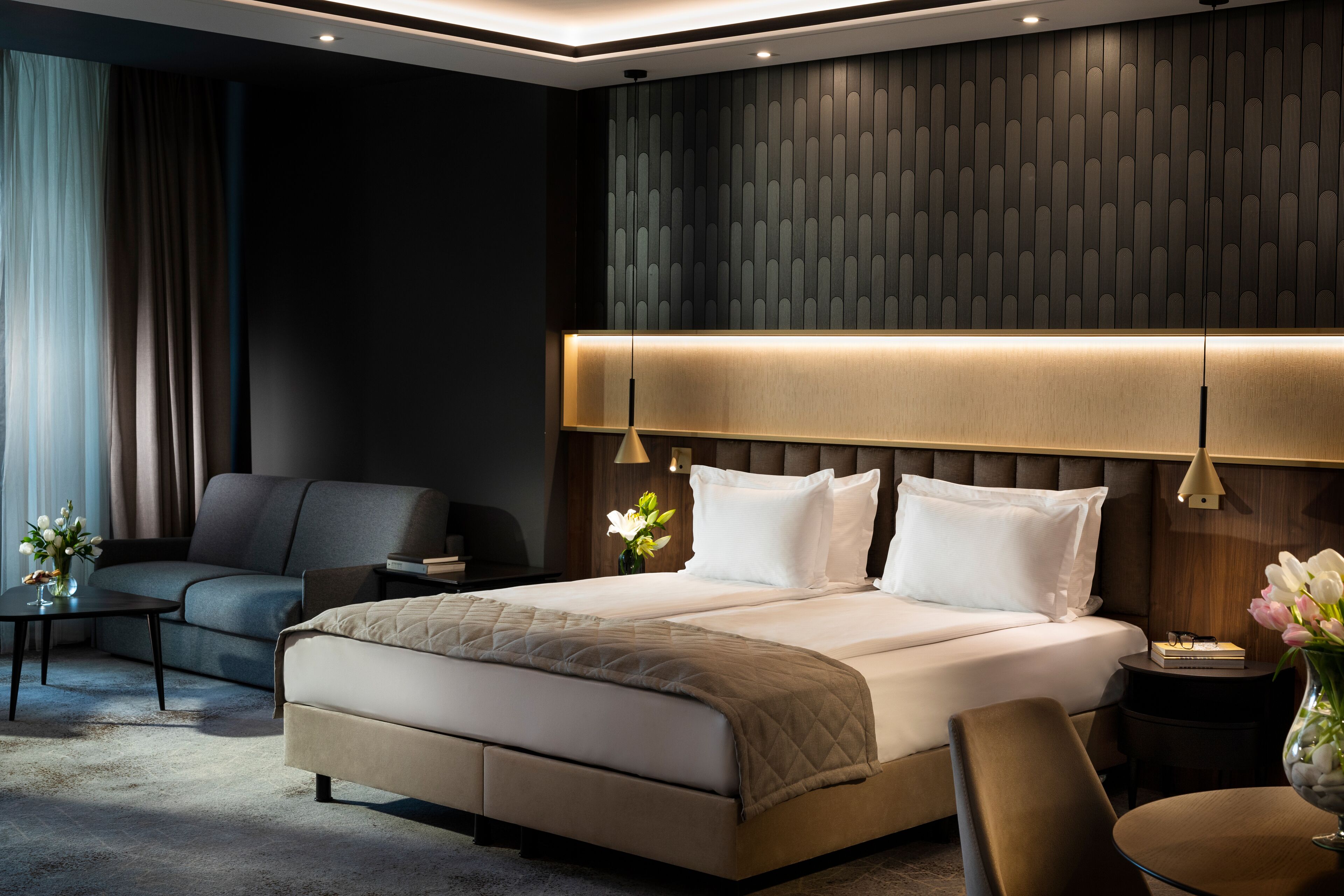 A modern hotel room with a large bed, a sofa, a small table with a vase of flowers, and a nightstand with a lamp.