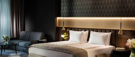 A modern hotel room with a large bed, a sofa, a small table with a vase of flowers, and a nightstand with a lamp.