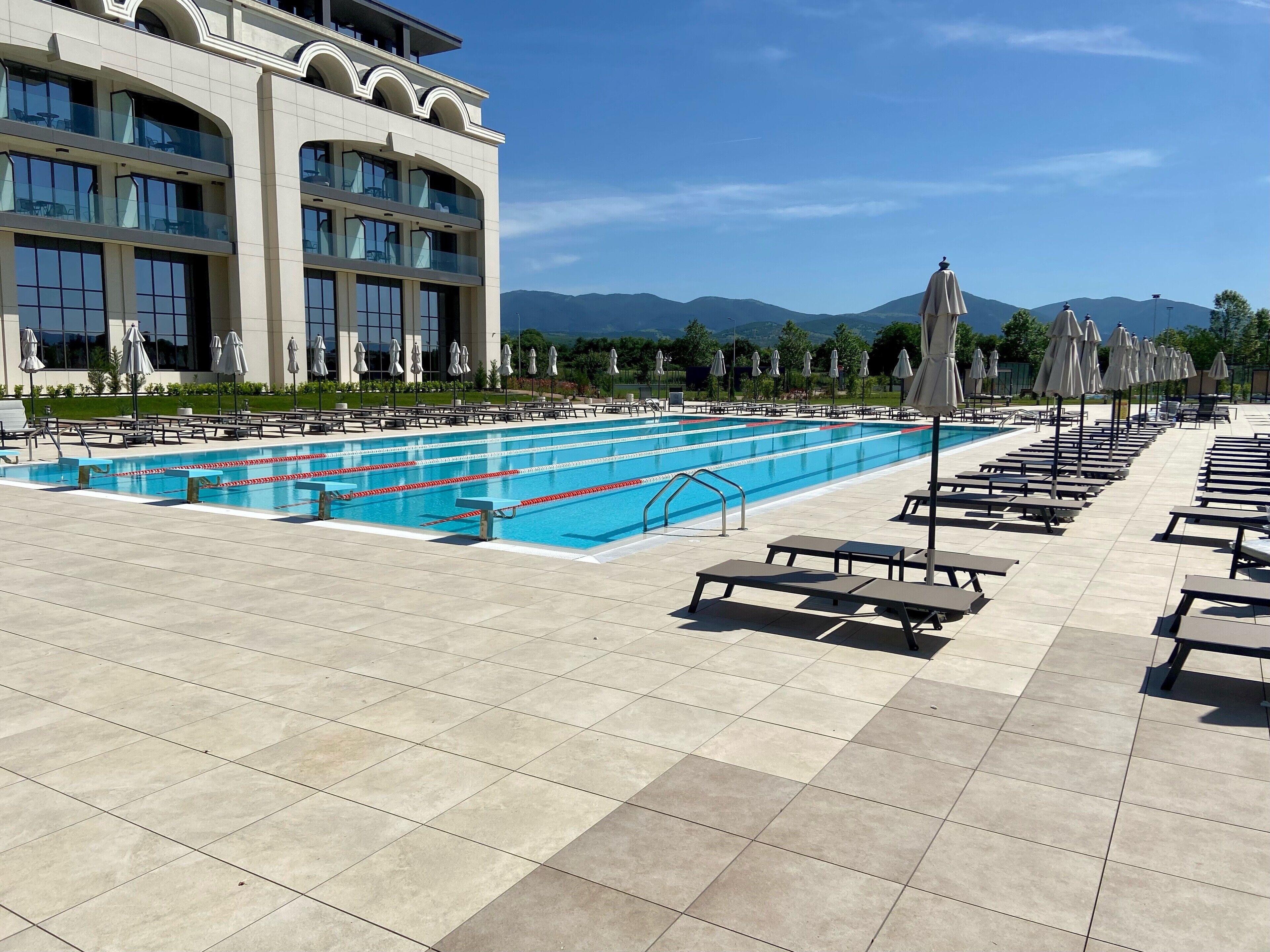 Une piscine entourée de chaises longues et de parasols, bordée d’un bâtiment aux grandes fenêtres, avec une chaîne de montagnes en toile de fond.