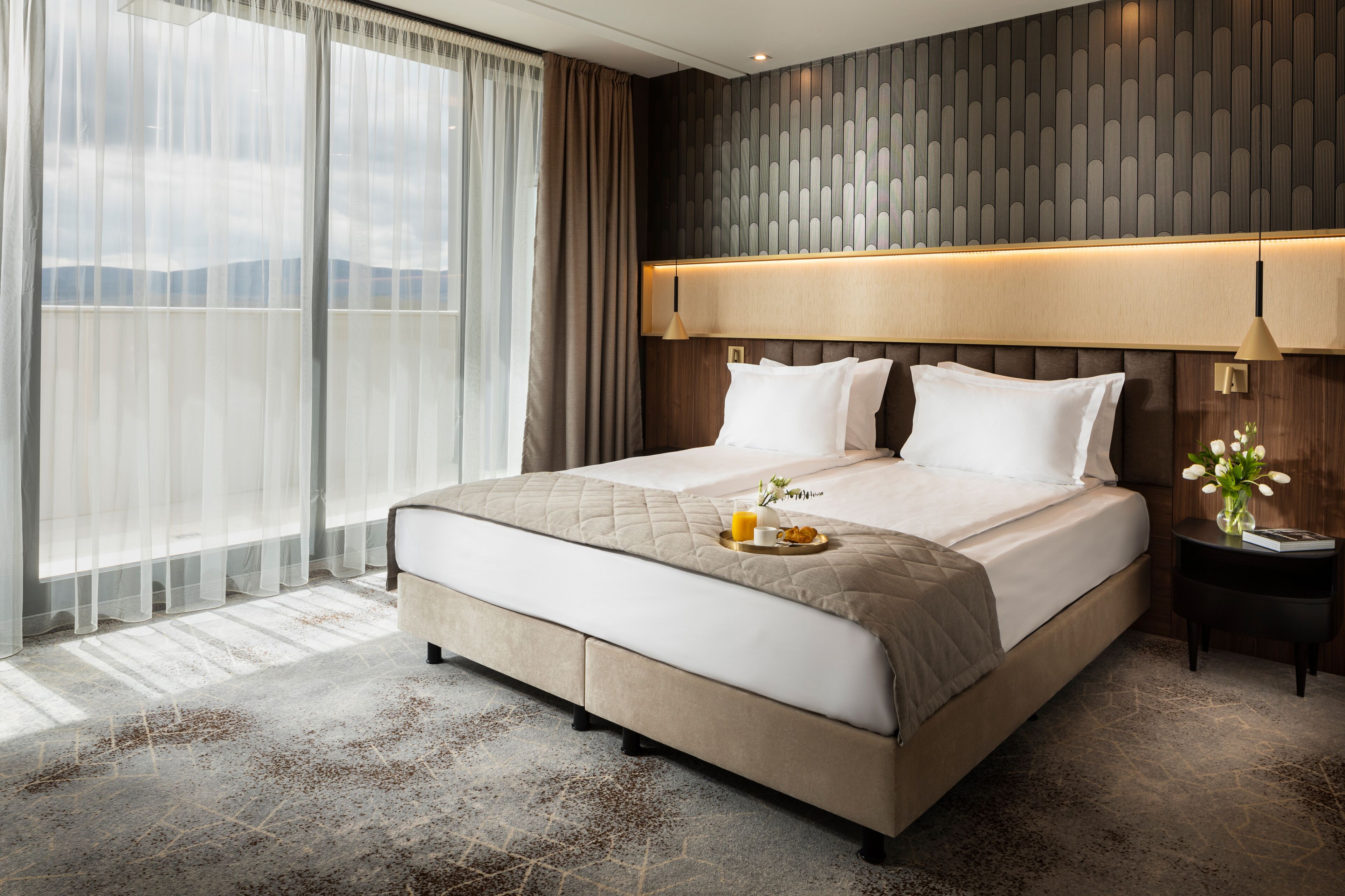A large bed with a quilted headboard, a bedside table with a lamp, and a view of mountains through sheer curtains.