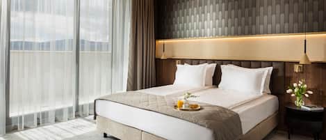 A large bed with a quilted headboard, a bedside table with a lamp, and a view of mountains through sheer curtains.