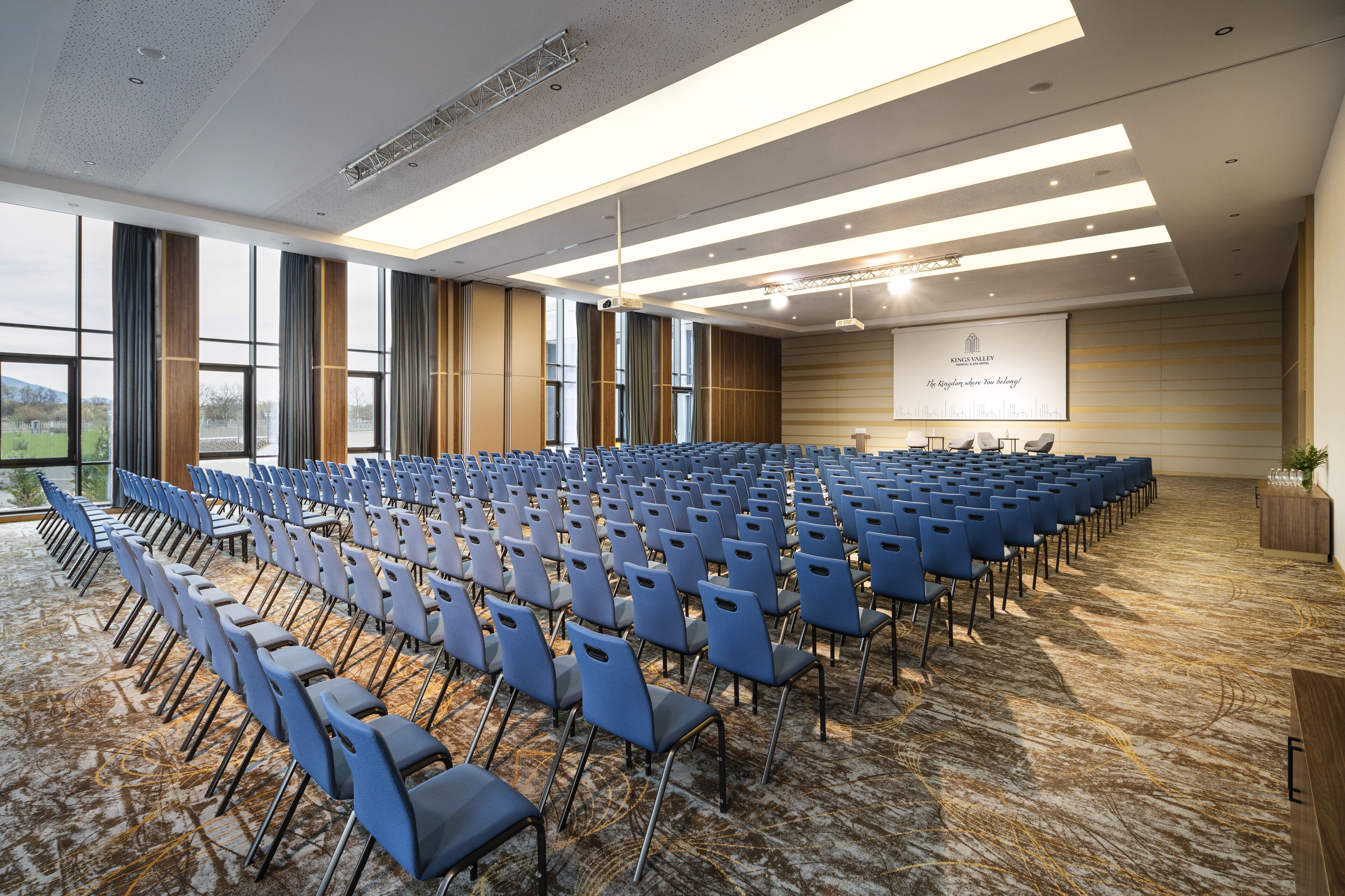 Une salle de conférence avec des rangées de chaises bleues, un écran de projection et une grande fenêtre donnant sur l’extérieur.