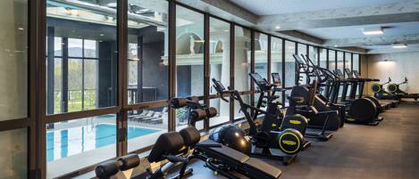 A gym with various exercise equipment, large windows, and a pool visible through the windows.