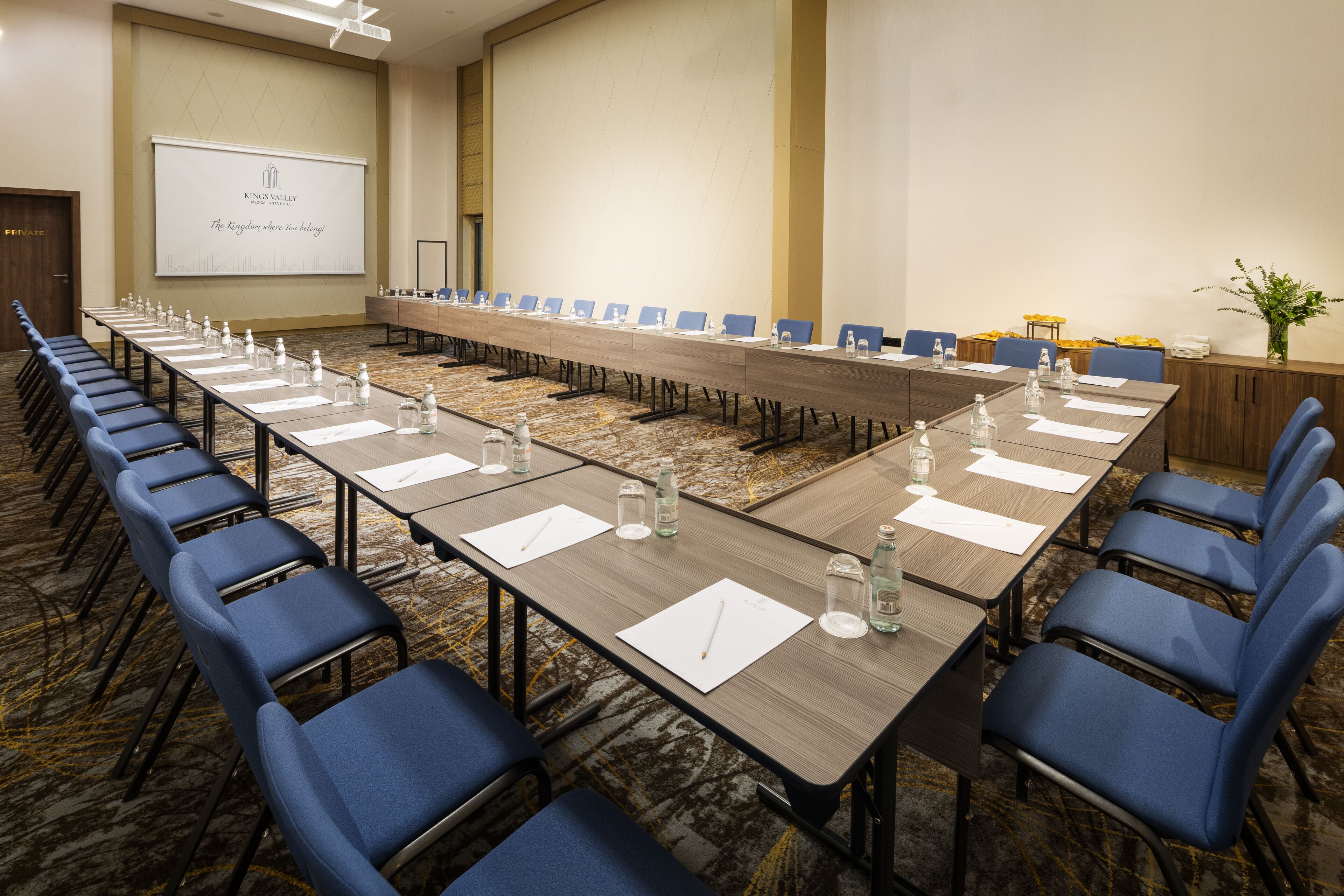 A conference room with a long table, blue chairs, water bottles, and a projector screen displaying "King Valley The King where you belong!"