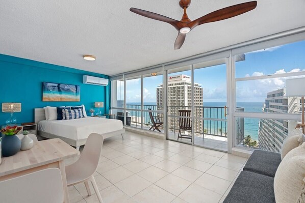 A modern hotel room with a bed, a desk, and a balcony with a view of the city and ocean. - Imperial Suite | Spacious studio with stunning ocean views! (San Juan)