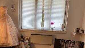 Huoneessa on suuri ikkuna, patteri, vaaleanpunainen jalkarahi, tiikurikuvamaalaus ja tuolilla oleva mekko.