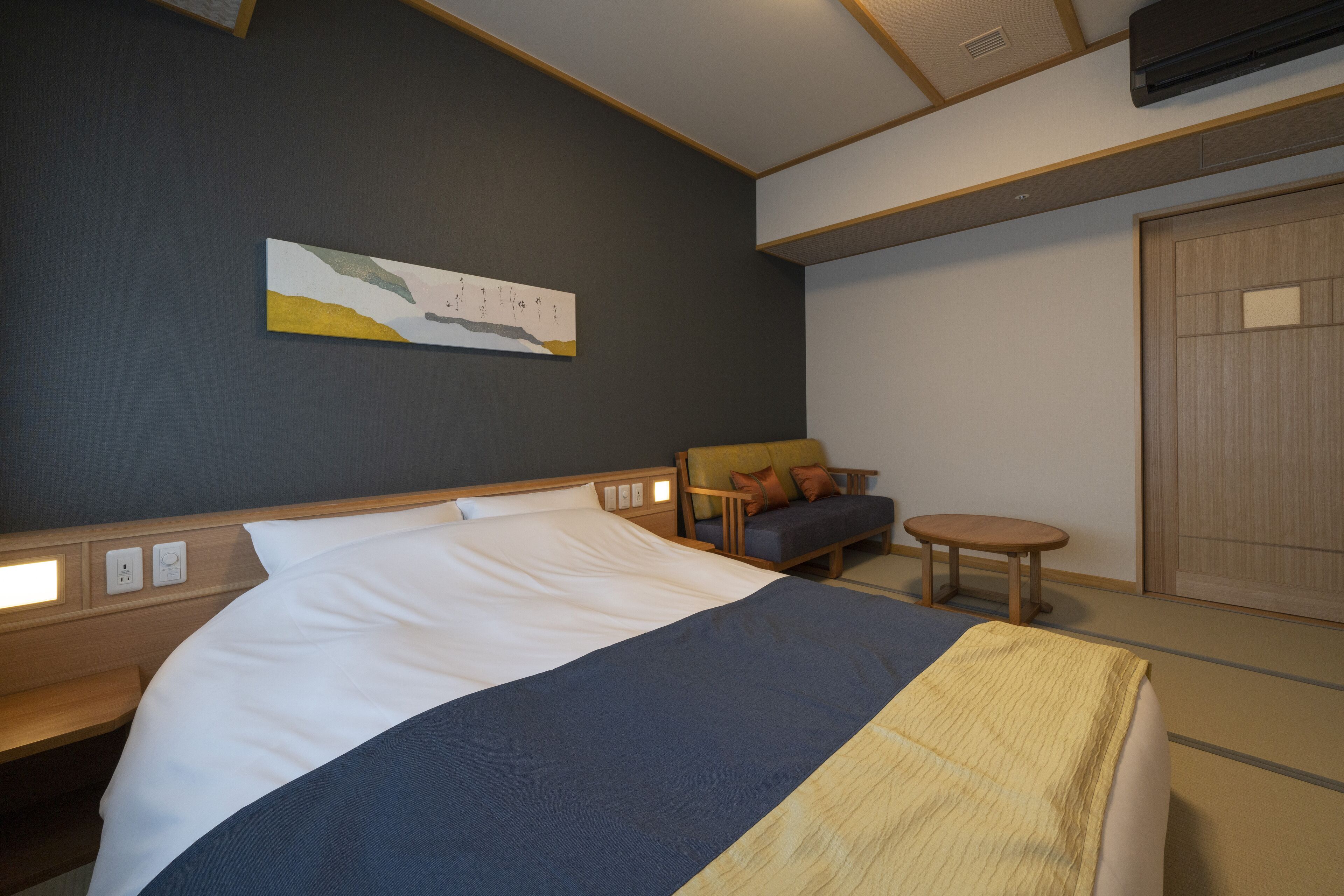 A hotel room with a bed, a small sofa, a round table, and a wall-mounted artwork.