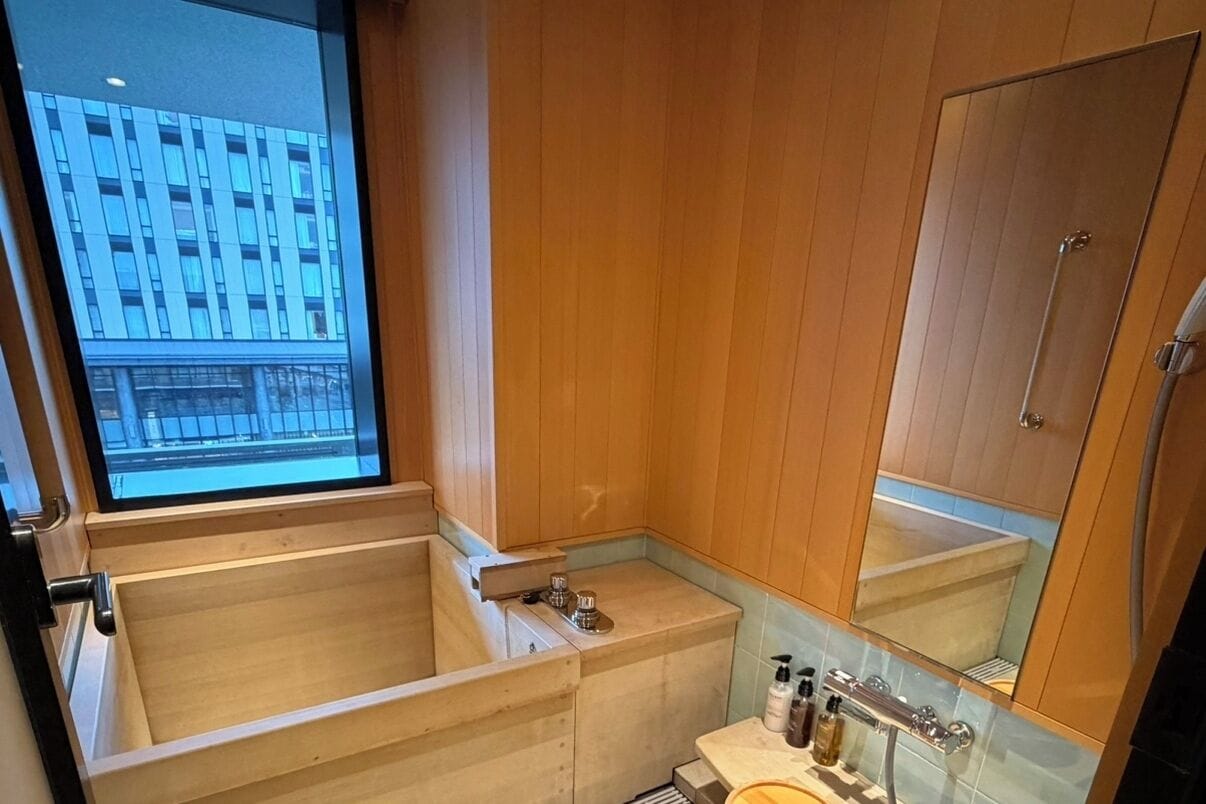 A modern bathroom with a wooden vanity, a large bathtub, and a window with a view of a building.