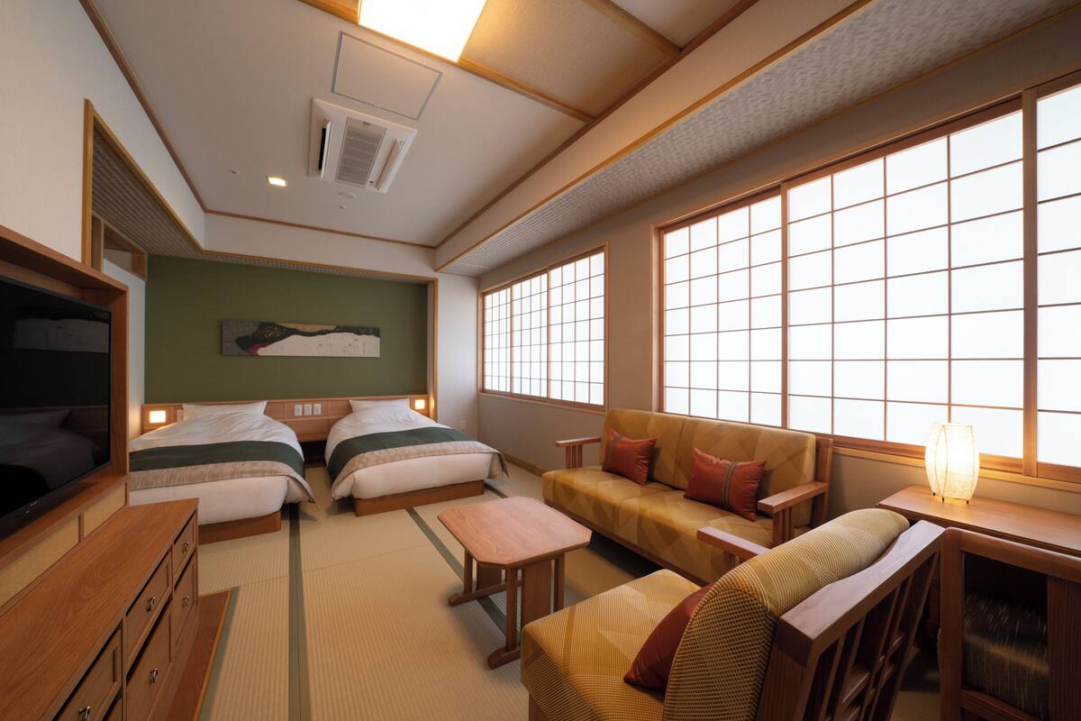 A traditional Japanese-style room with tatami flooring, a futon bed, a low wooden table, a cushioned sofa, and a large window with shoji screens.