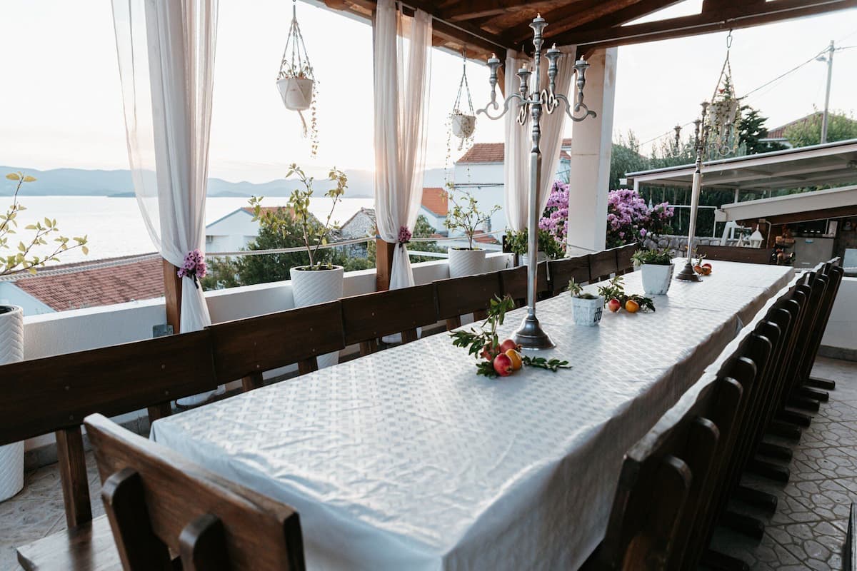 An outdoor dining area with a long table, chairs, hanging plants, and a view of the sea.