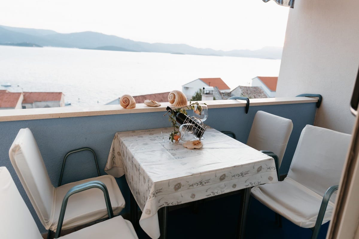 A small table with a floral tablecloth, surrounded by white chairs, overlooking a body of water with buildings in the distance.