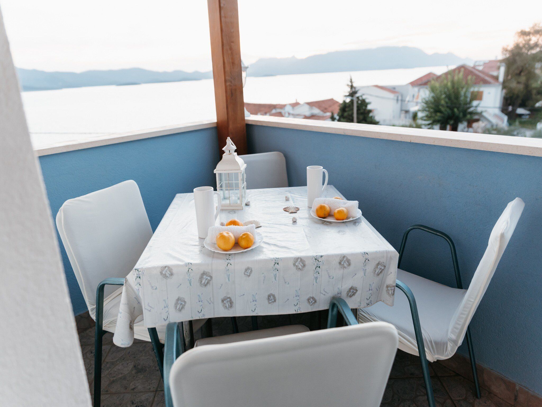 Un balcon avec une table dressée pour deux personnes, agrémenté d’une nappe blanche, de deux chaises, d’une lanterne blanche, d’une assiette d’oranges et d’une tasse blanche.