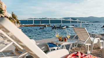 A beachside view with lounge chairs, a table with drinks and fruit, and a bridge in the background.
