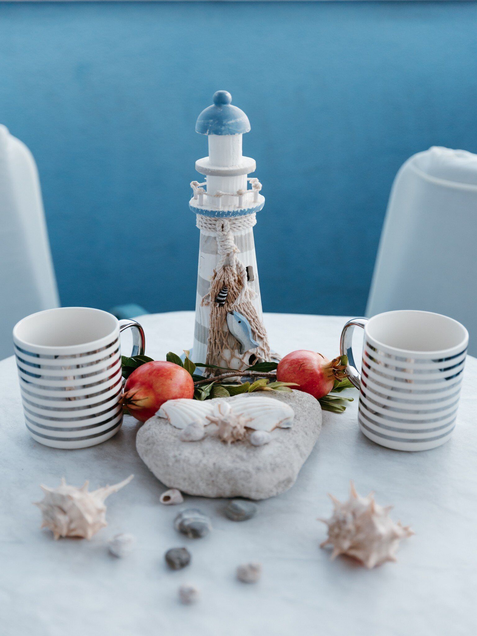 A table with a decorative lighthouse centerpiece, surrounded by seashells and pomegranates, with two striped mugs on either side.