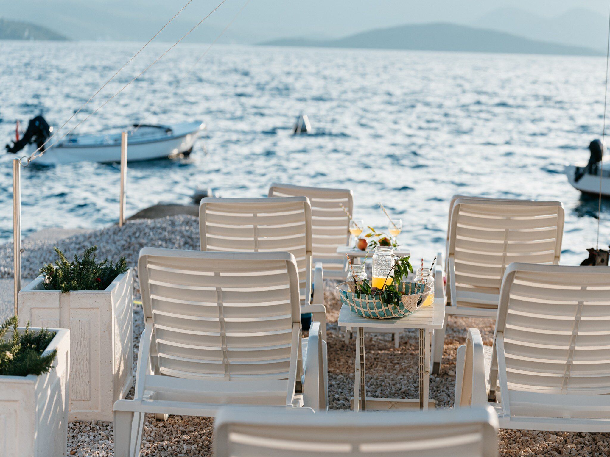 Ein Sitzbereich am Strand mit weißen Stühlen und Tischen, ein kleiner Tisch mit Getränken und einem Korb, sowie Boote im Hintergrund.