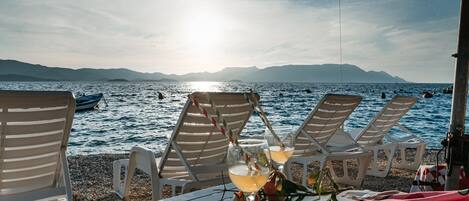 Ein Sitzbereich am Strand mit Liegestühlen und einem Tisch, mit Blick auf ein ruhiges Meer mit Booten und entfernten Bergen.