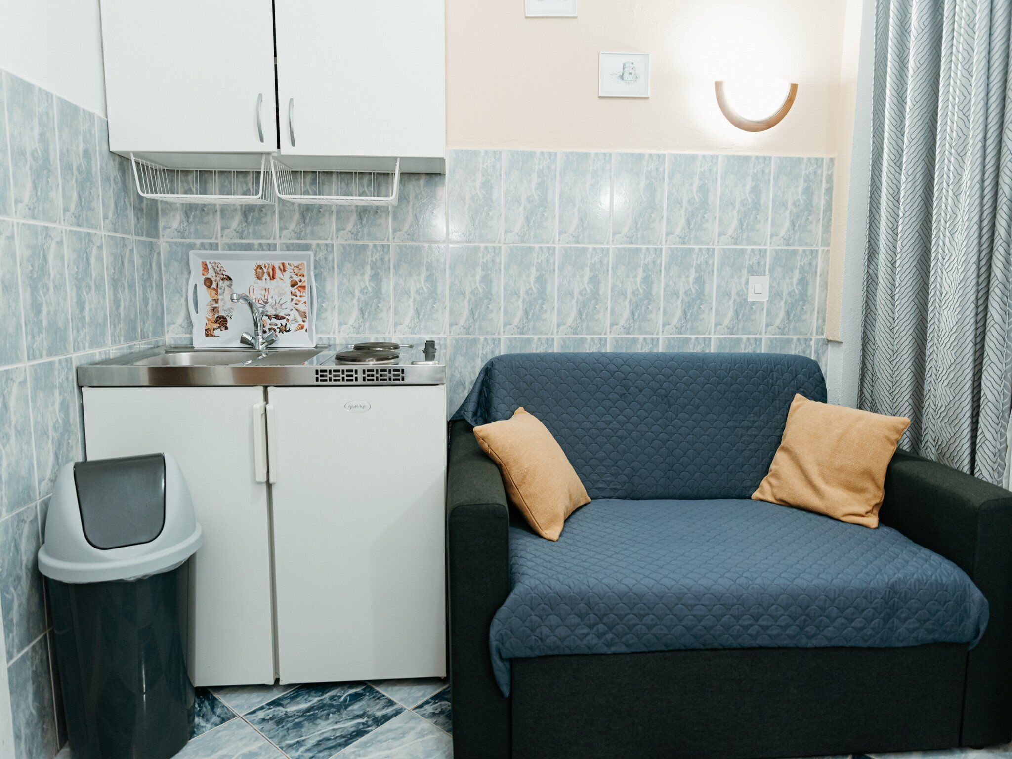 Une petite cuisine équipée comprenant un réfrigérateur, un évier et un four à micro-ondes. On y trouve une poubelle et une petite étagère avec des objets décoratifs. Un canapé bleu avec deux coussins est placé contre le mur.